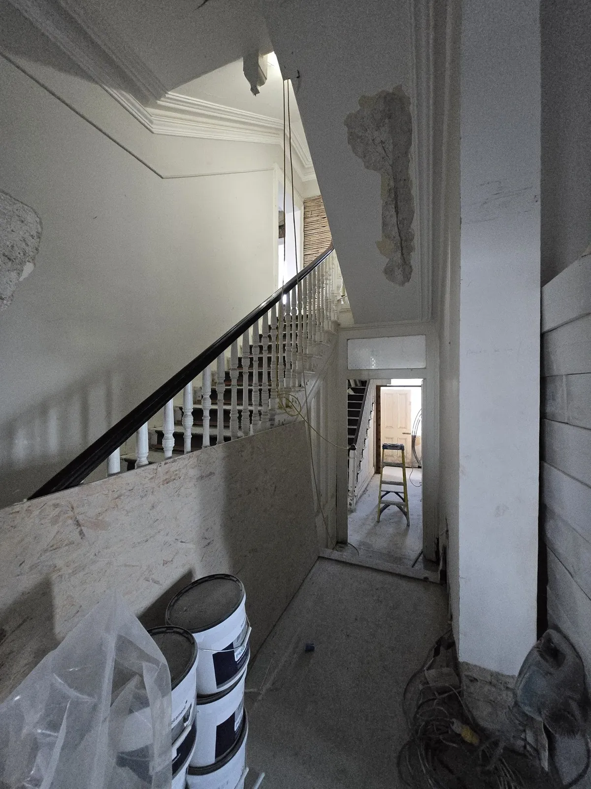 Stripped hallway of a protected period house mid-renovation, with failed plaster patches above the door, bare walls and dust sheets beside the stairs